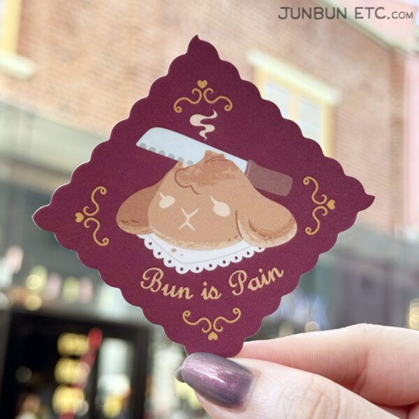 A photo showing a sticker design with JunBun's head as a bread boule, being sliced into by a bread knife, with gentle steam rising from it, with curlicue border brackets. The serif font below states "Bun is Pain."