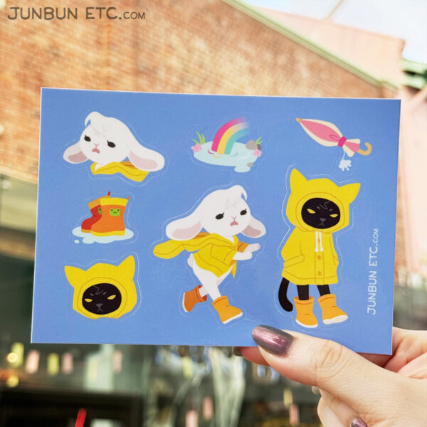 The graphic shows the November Rain sticker sheet image depicting JunBun - the white rabbit mascot - fleeing the rain with the hood of their raincoat waving in the wind, and MaxCat - the black cat mascot - togged up fully in his raincoat, listlessly kicking puddles with his galoshes. Alongside them are stickers of a pair of wellies in a puddle with frog head detailing on them, a rainbow coming out of a puddle, and a closed umbrella with a raincloud charm.