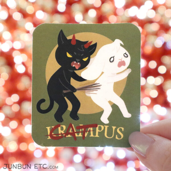 A photo showing the Catmpus single sticker design with MaxCat as Krampus with horns and an extended tongue, catching a despairing JunBun and switching them as per Krampus tradition. The title below, "Krampus," is partially cancelled out and "Cat" is written over the start of the word.