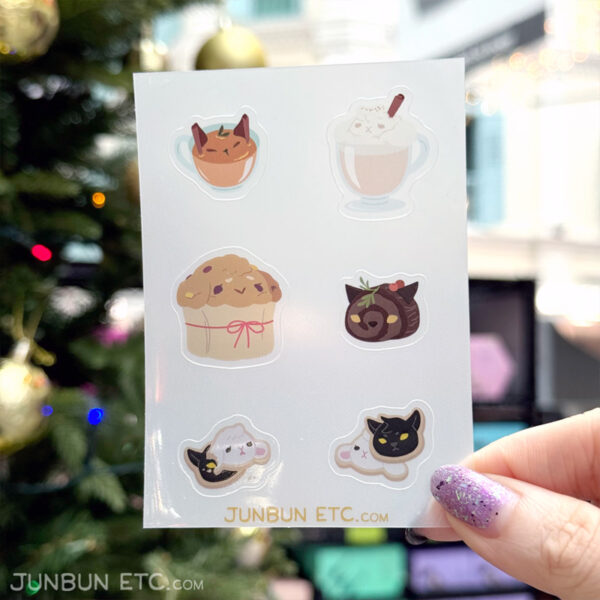 A photo showing a sticker sheet design of various seasonal holiday food that resemble JunBun and MaxCat. JunBun-themed foods include a glass of eggnog and a panettone, MaxCat-themed foods include a cup of spiced cider and a yule log slice. Sugar cookies resembling both of them are stacked on top of each other for the last 2 stickers - the one with MaxCat below has JunBun's cookie looking dismayed as a bite has been taken out of their ear.