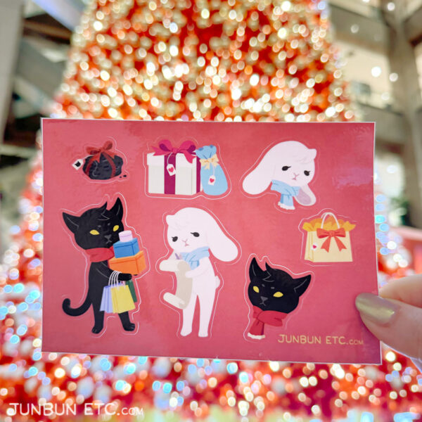 The photo shows the Naughty or Nice sticker sheet depicting JunBun - the white rabbit mascot - ticking off a long giftlist, and MaxCat - the black cat mascot - carrying bags and boxes of holiday gifts after holiday shopping, alongside colourful and cute wrapped Christmas presents, a gift bag, and a be-ribboned lump of coal.