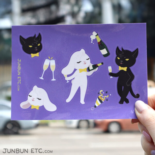 The photo shows the Champagne Toast sticker sheet depicting JunBun - the white rabbit mascot - holding a champagne glass and offering a champagne bottle tipple to MaxCat - the black cat mascot - jauntily leaning back and holding out his glass, alongside a bubbly bottle of champagne, two clinking glasses, and a party popper with emergent confetti.