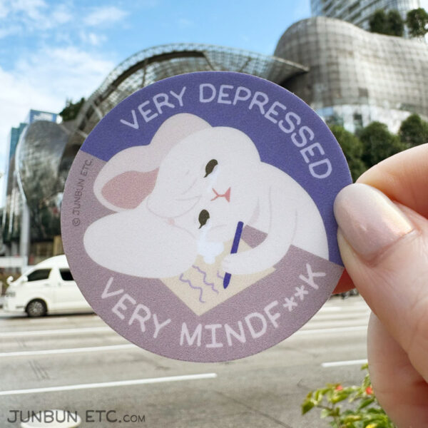 A photo showing the Very Depressed single sticker of JunBun - the white bunny mascot - stopping mid-writing, resting their head on the table weeping. Their ears have drooped onto the table and their tears are pooling. Above and below the main image is the caption: "very depressed, very mindf**k."