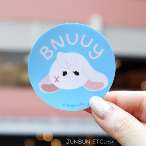 A photo showing the Bnuuy single sticker of JunBun's - the white bunny mascot - head with a neutral expression, one eye and nose swapped so that their nose starts first on their face, followed by two eyes. Above the head is an arched "BNUUY" in rounded and simple block handwriting.