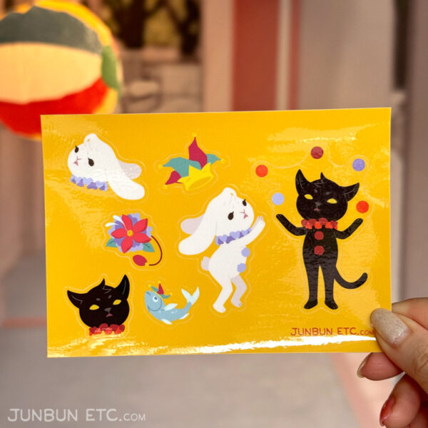 The photo shows the Clowning Around sticker sheet depicting JunBun - the white rabbit mascot - and MaxCat - the black cat mascot - as clowns in a circus. MaxCat is juggling five balls with a sage expression, and a rightfully-concerned JunBun hovering nearby is reaching to catch a ball veering off-course. Alongside that are a four-pointed jester hat complete with bells on the hat-ends, a lapel flower squirting water, and a flopping fish with a little jester hat.