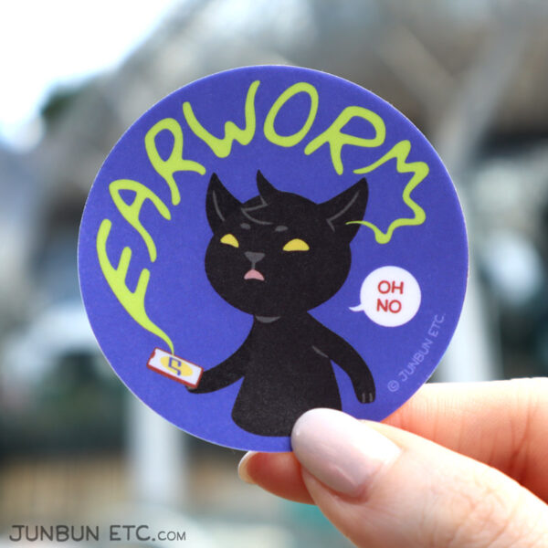A photo showing the Earworm single sticker of MaxCat - the black cat mascot - holding a phone with music playing on its screen, looking up at the word "Earworm" in wiggly handwritten font emerging from the phone and snaking towards his ear. He succinctly says "oh no," in a speech bubble next to him.