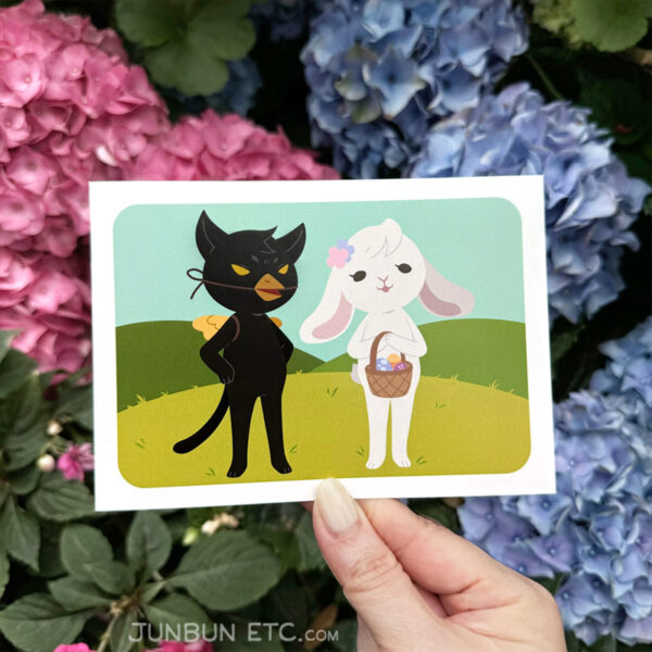 The graphic shows the Egg Hunt postcard image depicting JunBun - the white rabbit mascot - as the Easter rabbit, beatifically holding a basket of painted eggs and flowers on their head. MaxCat - the black cat mascot, is dressed as a chick, replete with fake beak and wings, looking right pissed off. They stand in a peaceful pastoral scene of spring-green hills.