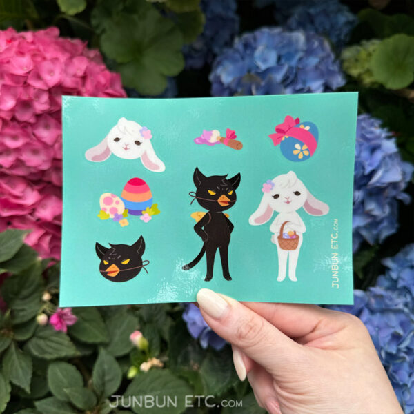 The photo shows the Egg Hunt sticker sheet image depicting JunBun - the white rabbit mascot - as the Easter rabbit, beatifically holding a basket of painted eggs and flowers on their head. MaxCat - the black cat mascot, is dressed as a chick, replete with fake beak and wings, looking right pissed off. Alongside that are a blue and yellow painted egg with a sweet red ribbon tied into a bow around it, a bow-adorned paintbrush with purple paint, and a pair of eggs placed in flowers and grass.