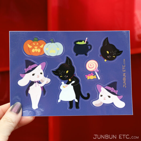 The photo shows the CatWitchween sticker sheet depicting JunBun - the white rabbit mascot - dressed up in a witch costume cheerily holding a wand, and MaxCat - the black cat mascot - dressed up as a familiar with a bell collar, checking out their Treat-or-Treating spoils, along with a bubbling cauldron drink, a lollipop with two pieces of candy, and a set of Jack-o-Lantern pumpkins - one larger and orange with a scary face, and the other pale teal with a terrified face.