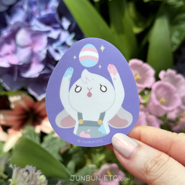 A photo showing the Egg single sticker depicting JunBun - the white bunny mascot - in an blue and pink paint-splattered apron, paws similarly splattered, enraptured by a sparkling egg painted in blue, pink and white bands above their head. The egg is painted the transgender pride flag.