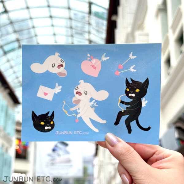 The photo shows the Loveshot sticker sheet depicting JunBun - the white rabbit mascot - and MaxCat - the black cat mascot - as Valentine's Cupids, wherein a grimacing MaxCat has accidentally shot an agonised JunBun with an arrow. Alongside that are a pair of winged Cupid arrows with hearts as arrowheads, a winged letter sealed with a heart, and a squishy heart pierced by a winged arrow, with hearts flowing from the impact.