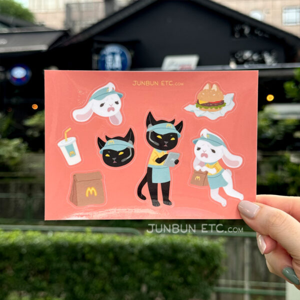 The photo shows the MewBunny's sticker sheet depicting JunBun - the white rabbit mascot - and MaxCat - the black cat mascot - as fast food/diner/burger joint minimum wage workers, complete in MewBunny's uniforms and visors. MaxCat is reviewing orders on a tablet, and JunBun is falling over in dismay with a brown paper bag in paw. Alongside that are a cat-eared unwrapped cheeseburger, a soda cup drink, and a brown paper bag with the MewBunny's logo.