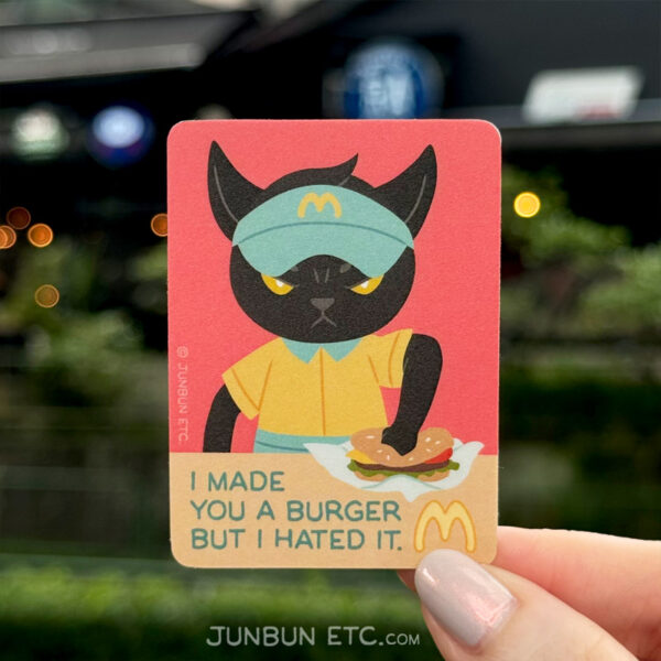 A photo showing the Smashed single sticker design depicting MaxCat - the black cat mascot - as a uniformed MewBunny's fast food worker, staring angrily at the viewer, a fist smashing a burger into the counter in front of him. The caption next to the MewBunny's logo says "I made you a burger but I hated it," in the vein of a classic derivative lolcat meme, "I made you a cookie but I eated it."